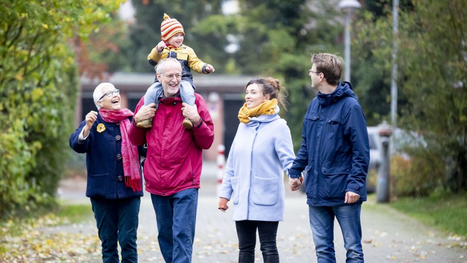 Junges Elternpaar und Großelternpaar beim gemeinsamen Spaziergang. Auf den Schultern des Großvaters sitzt ein lachendes Kleinkind.