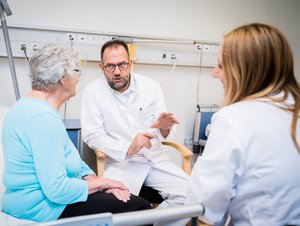 Eine ältere Frau sitzt in einem Krankenhausbett und spricht mit einem Arzt und einer Ärztin.